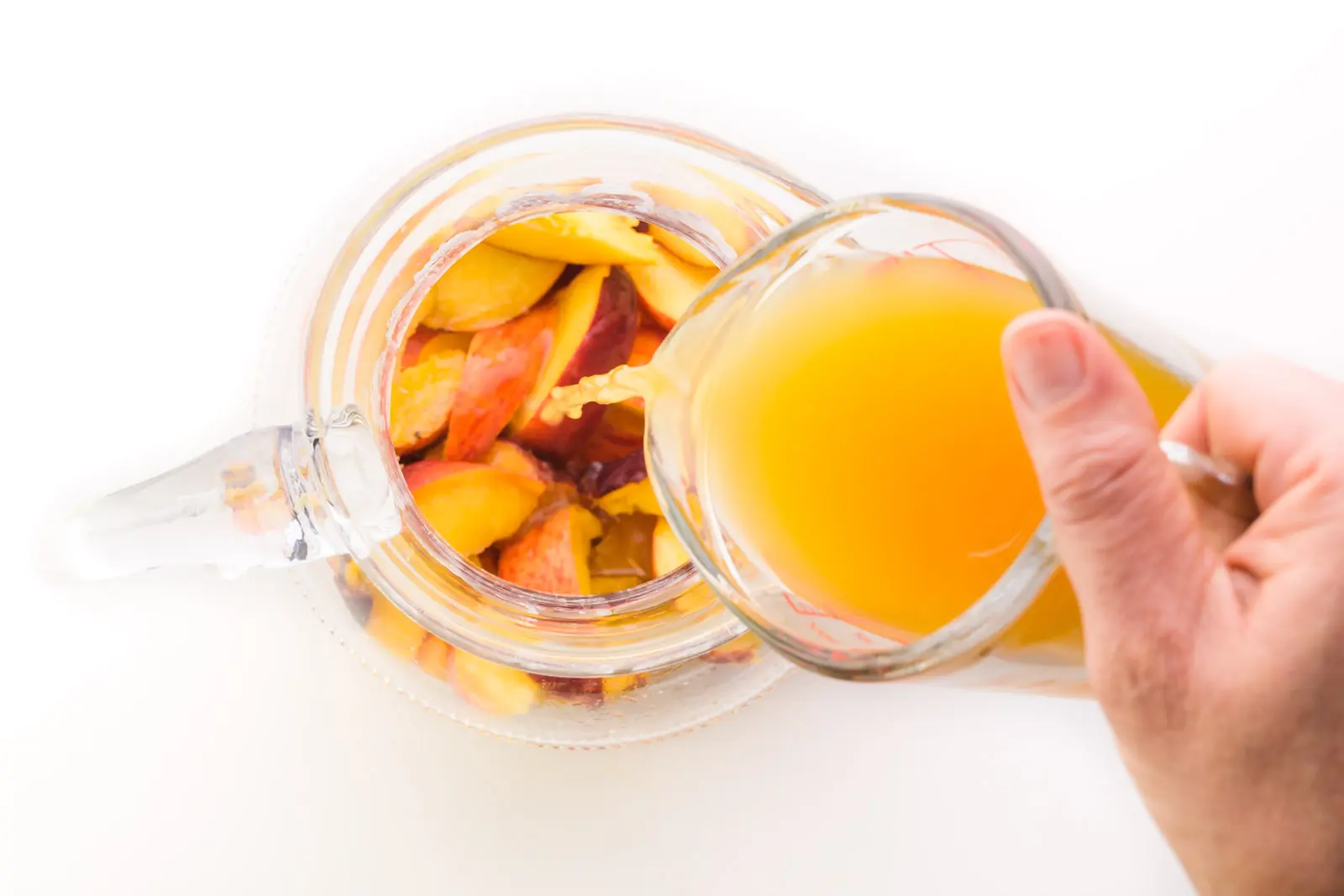 A hand holds a glass measuring cup full of peach juice and is pouring it into a pitcher with sliced peaches.