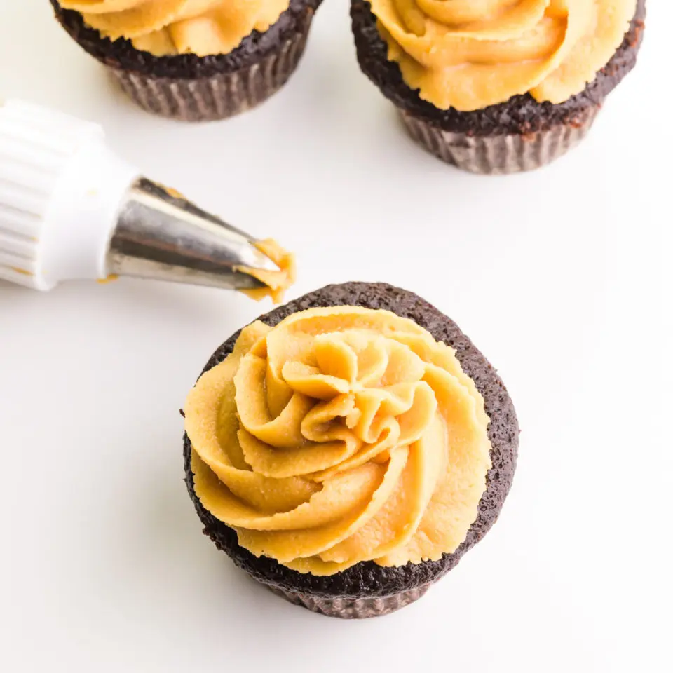 Looking down on three cupcakes with peanut butter frosting on top. The tip of a piping bag full of the frosting sits between them.