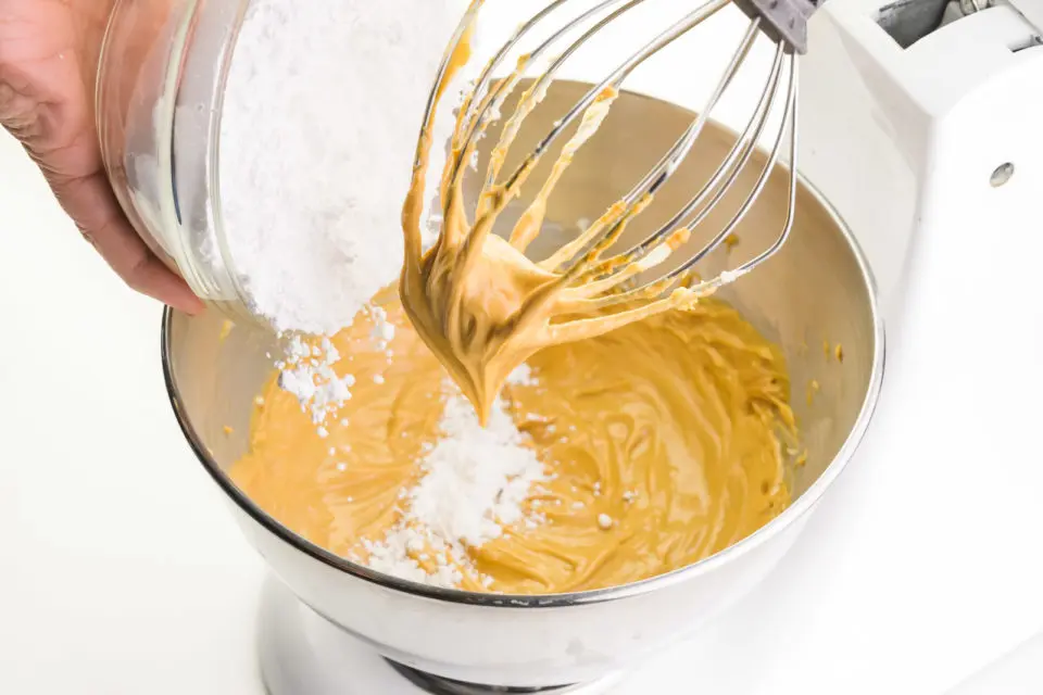 A hand holds a bowl of powdered sugar and is pouring it into a stand mixer with peanut butter mixture in the bottom and on the wire whisk beater.