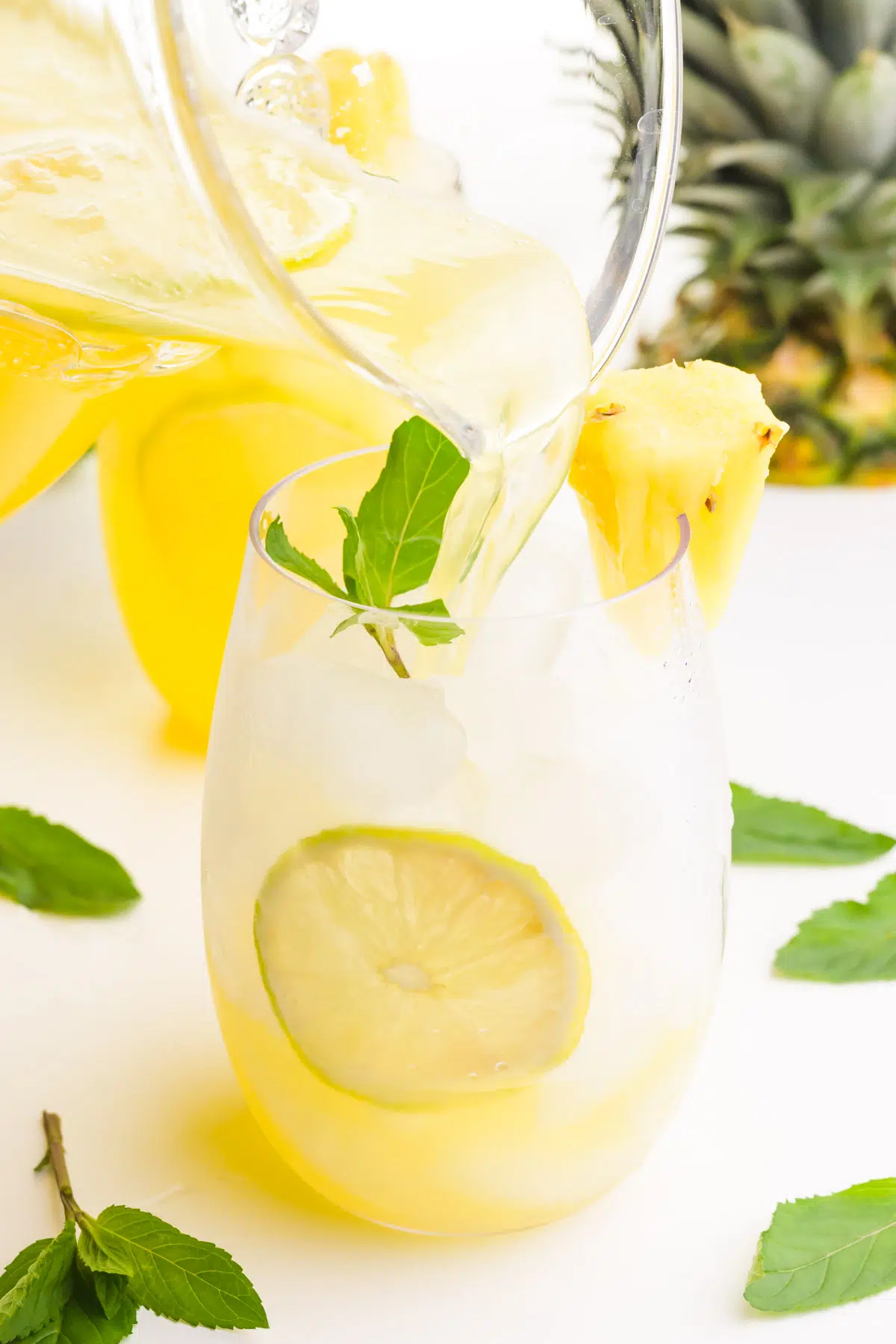 A pitcher is pouring pineapple beverage into a glass of ice with lime slices. There is a mint sprig and pineapple chunk on the glass. There are mint sprigs around the glass and the top of a fresh pineapple in the background.