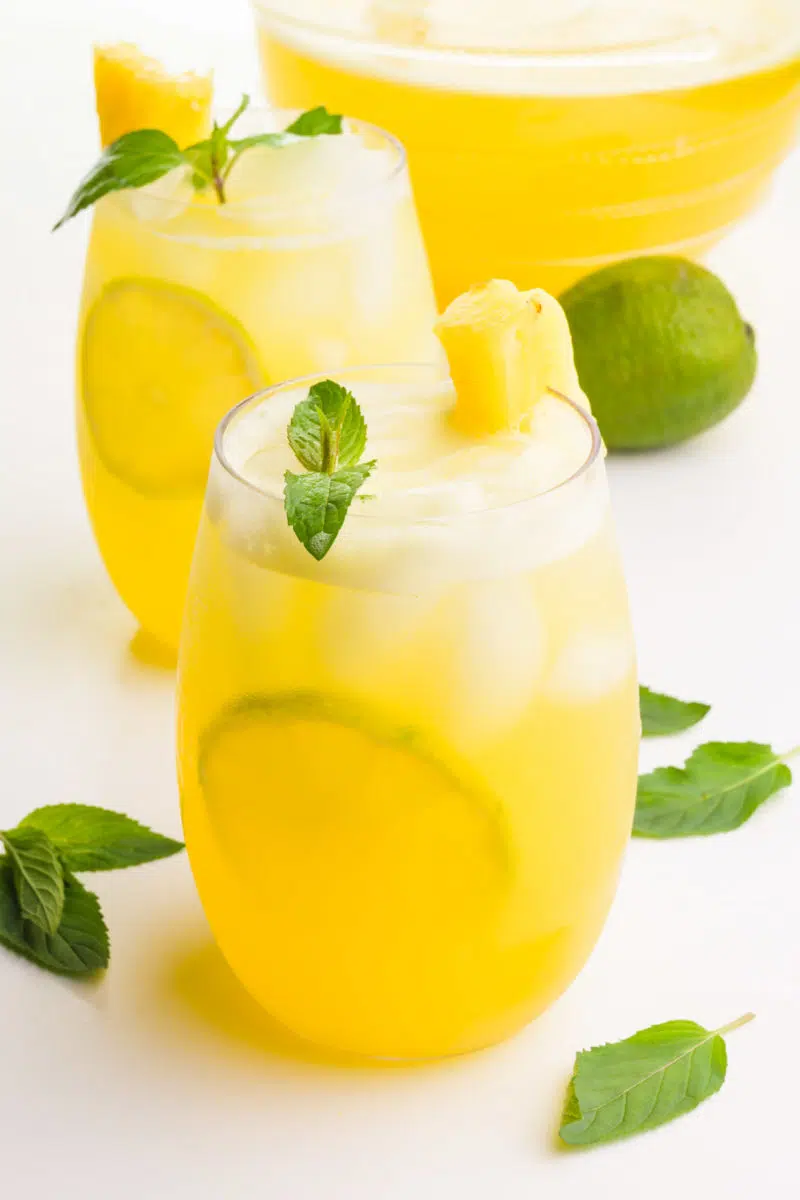 Two glasses sit one in front of the other. Both hold pineapple drinks with fresh limes, mint sprigs, and pineapple chunks. There are more mint herbs, a lime, and a pitcher with more of the beverage in the background.