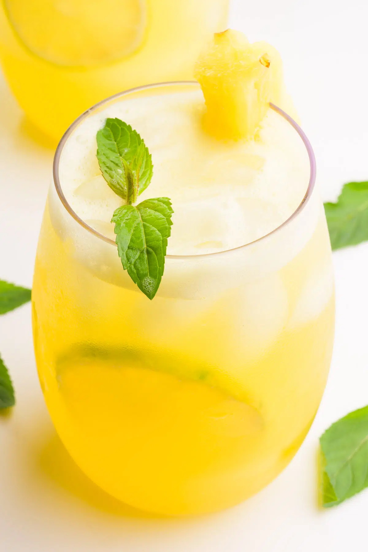 A glass holds pineapple agua Fresca with ice cubes and lime slices. There are mint sprigs in the glass and around it. There's another glass in the background.