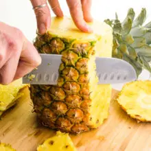 The top and bottom of a fresh pineapple have been chopped off. A hand holds a knife, cutting down the outer edge of the pineapple to remove the outer layer.