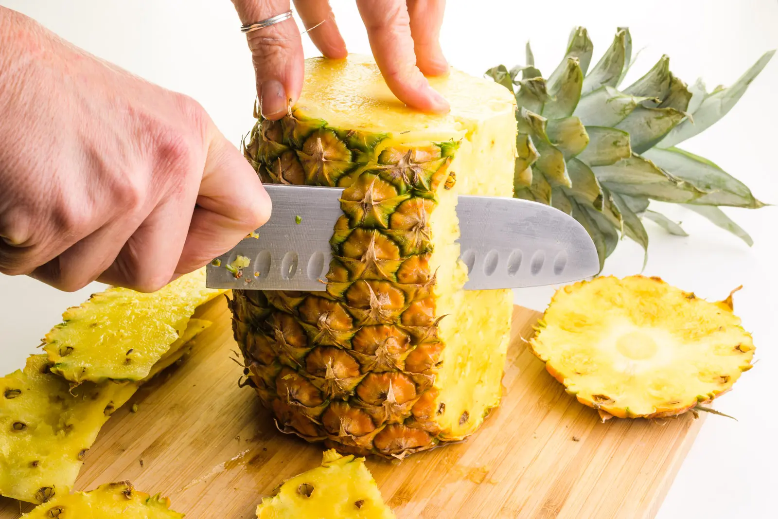 A hand holds a knife, cutting down the edge of a pineapple to remove the outer layer.
