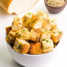 A bowl holds vegan croutons. There is a loaf of bread and a bowl of herbs barely visible in the background.