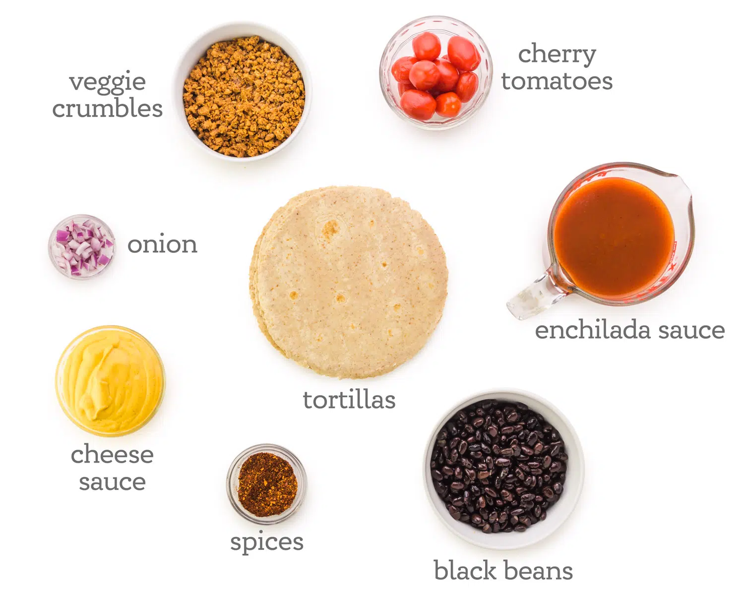 Ingredients are spread across a white table. The labels next to them read, cherry tomatoes, enchilada sauce, black beans, tortillas, spices, cheese sauce, onions, and veggie crumbles.