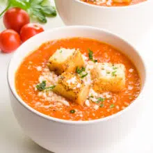 A bowl of tomato soup has croutons on top and fresh tomatoes in the background.