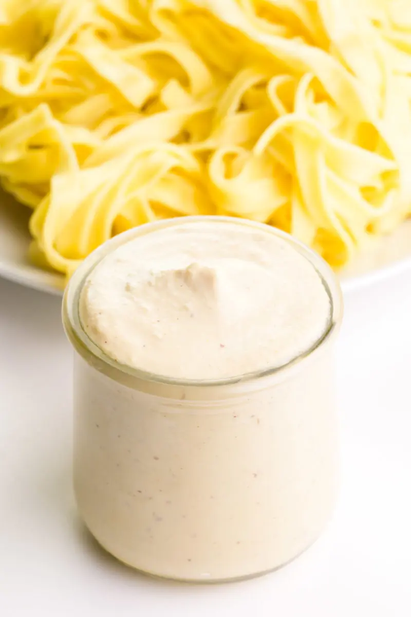 A small jar of vegan Alfredo Sauce sits in front of a plate full of cooked fettuccine noodles.