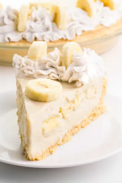 A slice of vegan banana cream pie sits on a plate. The rest of the pie sits behind it.