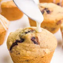A spoon hovers over a muffin, preparing to drizzle it with glaze. There are more muffins in the background.