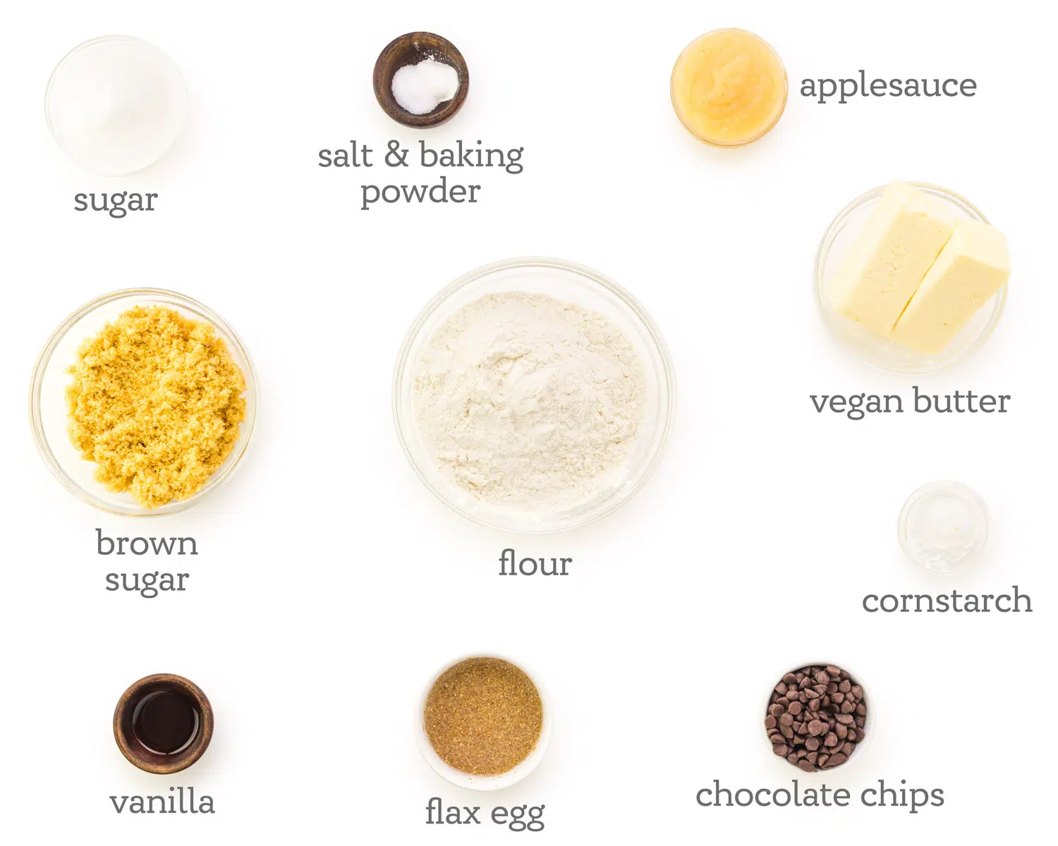 Ingredients are displayed on a counter. The labels next to them read, applesauce, vegan butter, cornstarch, chocolate chips, flax egg, vanilla, brown sugar, sugar, salt & baking powder, and flour.