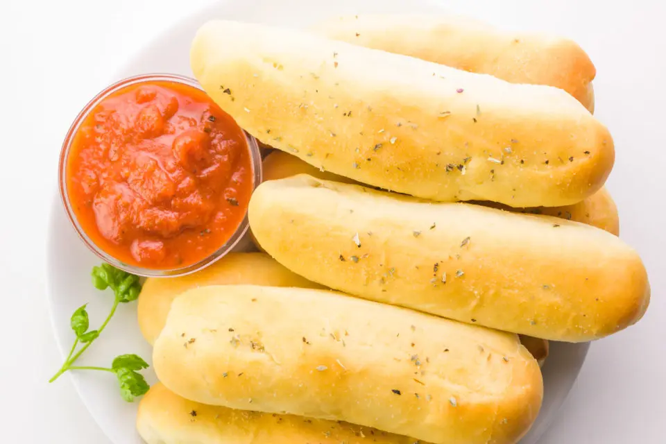 Garlic breadsticks are topped with herbs. They sit on a plate next to a bowl of marinara sauce and green herbs.