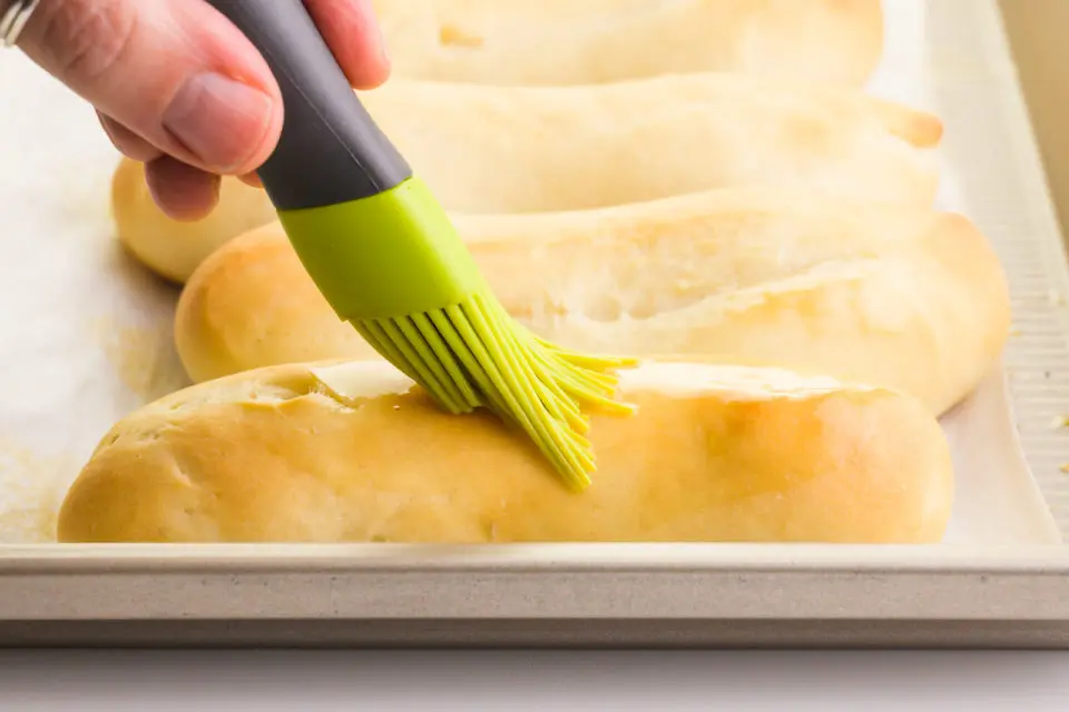 A hand holds a pastry brush, brushing melted butter over freshly baked garlic breadsticks.