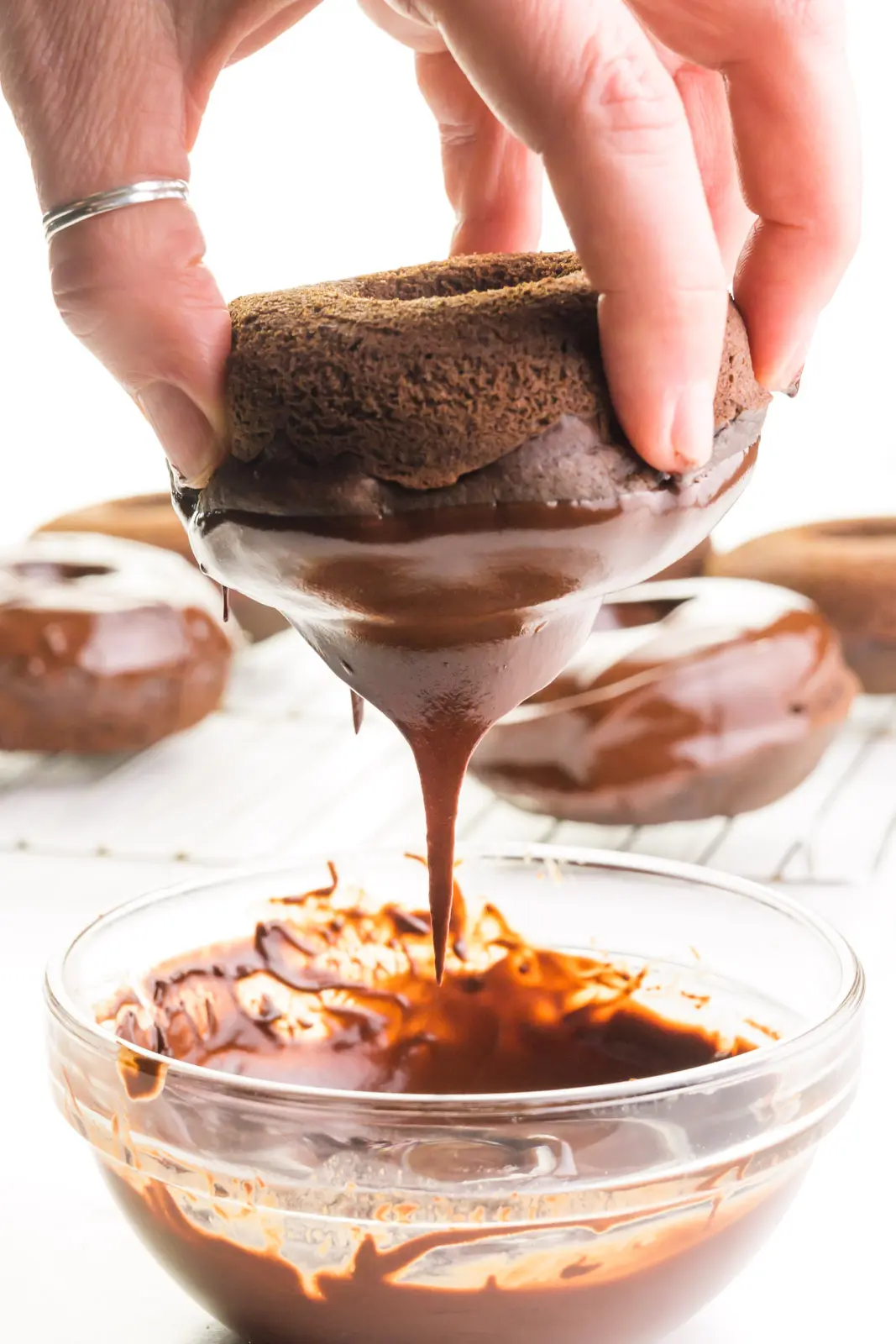 A hand holds a chocolate donut over a bowl of chocolate glaze. Some of the glaze is dripping off the donut. There are glazed donuts in the background.