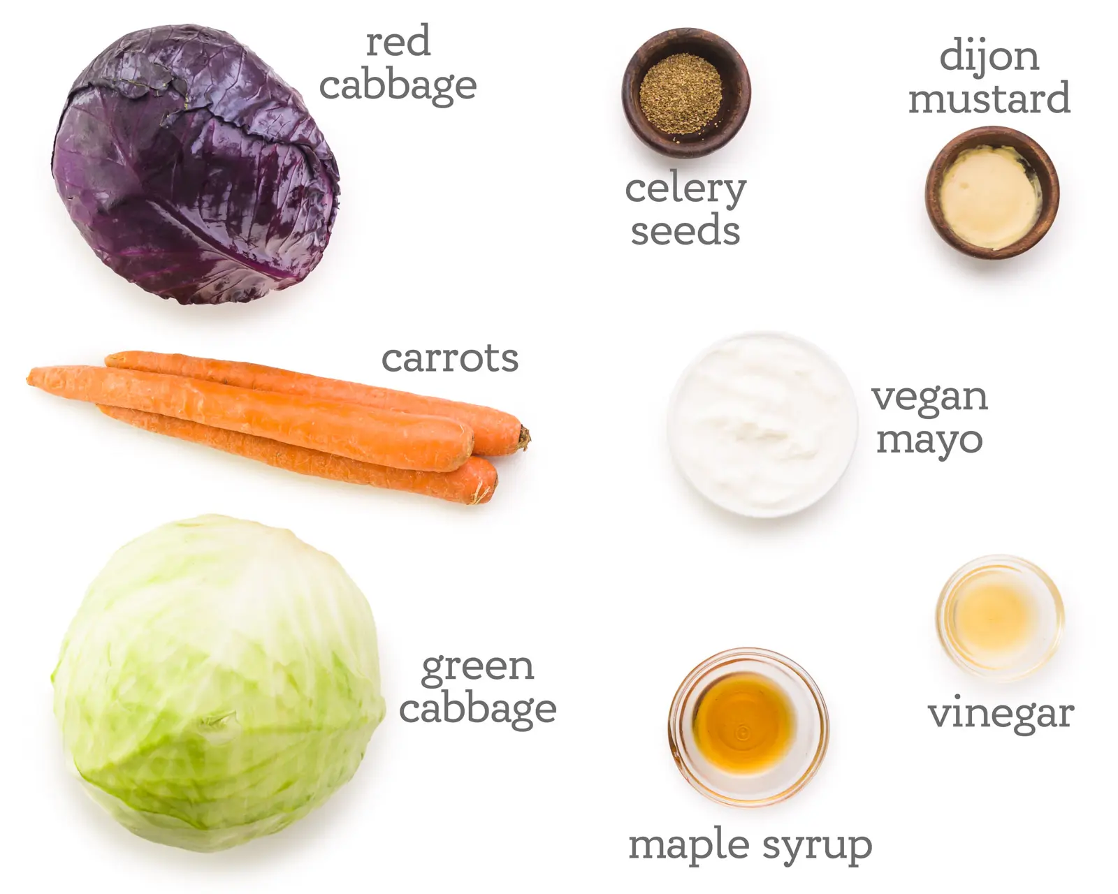 Ingredients are laid out on a table. The labels next to them read, celery seeds, dijon mustard, vegan mayo, vinegar, maple syrup, green cabbage, carrots, and red cabbage.