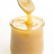 A spoon full of vegan condensed milk hovers over a glass jar full of the mixture.