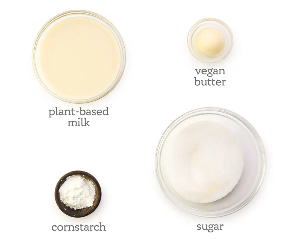 Ingredients in bowls are laid out on a white table. The labels next to them read, vegan butter, sugar, cornstarch, and plant-based milk.
