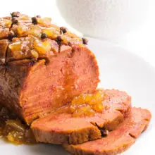 A vegan ham loaf has two slices cut out on a plate. There is pineapple sauce on top of the slices ad the loaf. There's a salad in the background.
