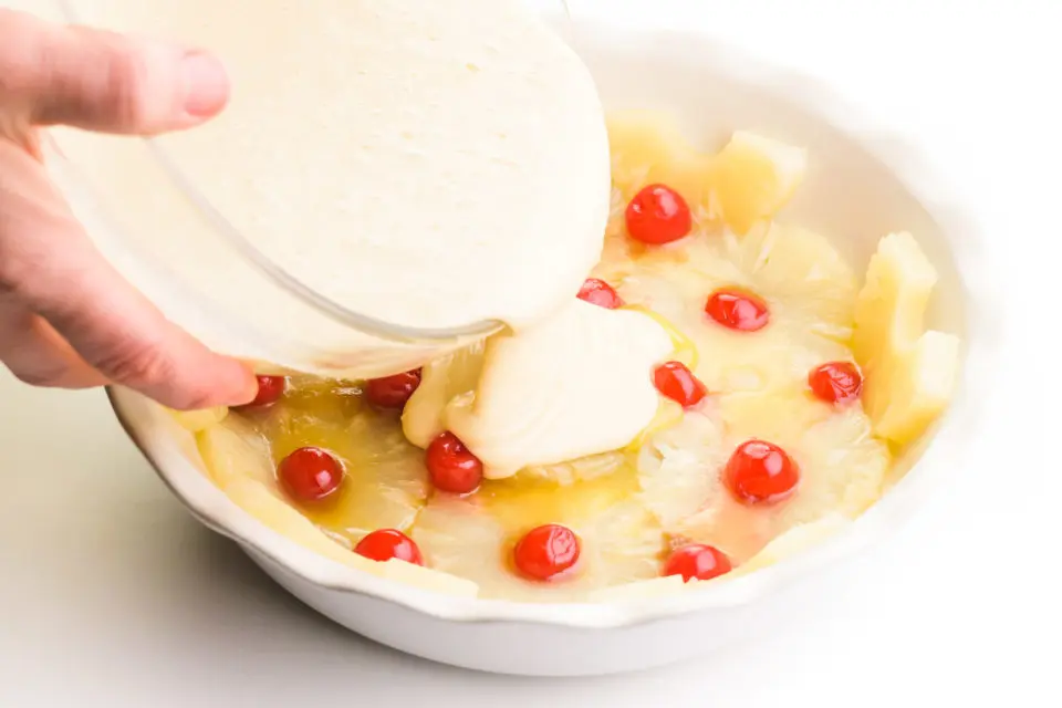 Cake batter is being poured over a pie pan lined with pineapple rings and cherries.