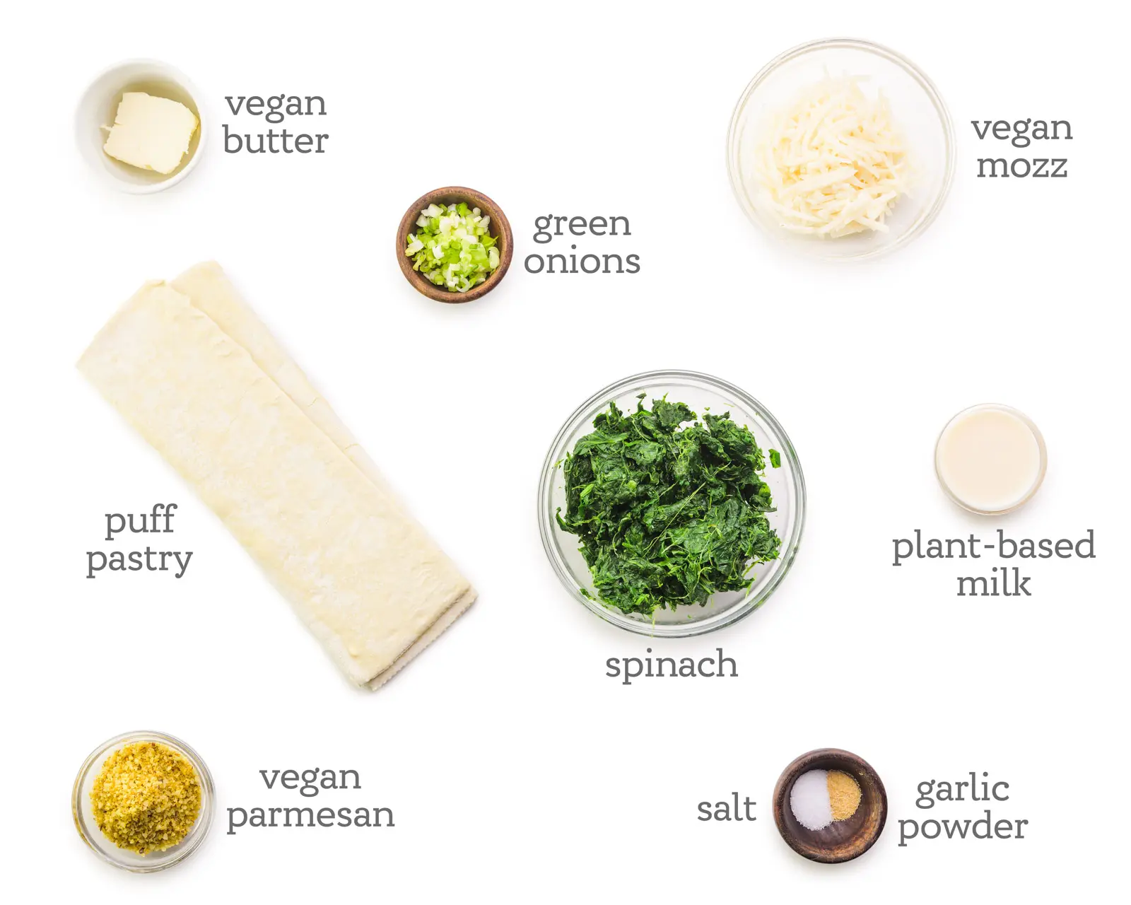 Ingredients are laid out on a white counter. The labels next to the ingredients read, vegan mozz, plant-based milk, garlic powder, salt, spinach, vegan parmesan, puff pastry, vegan butter, and green onions.