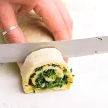 A hand holds a serrated knife, using it to cut slices out of a spinach pastry log.