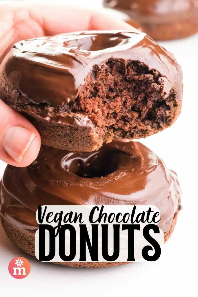 A hand holds a chocolate donut with a bite taken out, hovering over another one. The text reads, Vegan Chocolate Donuts.