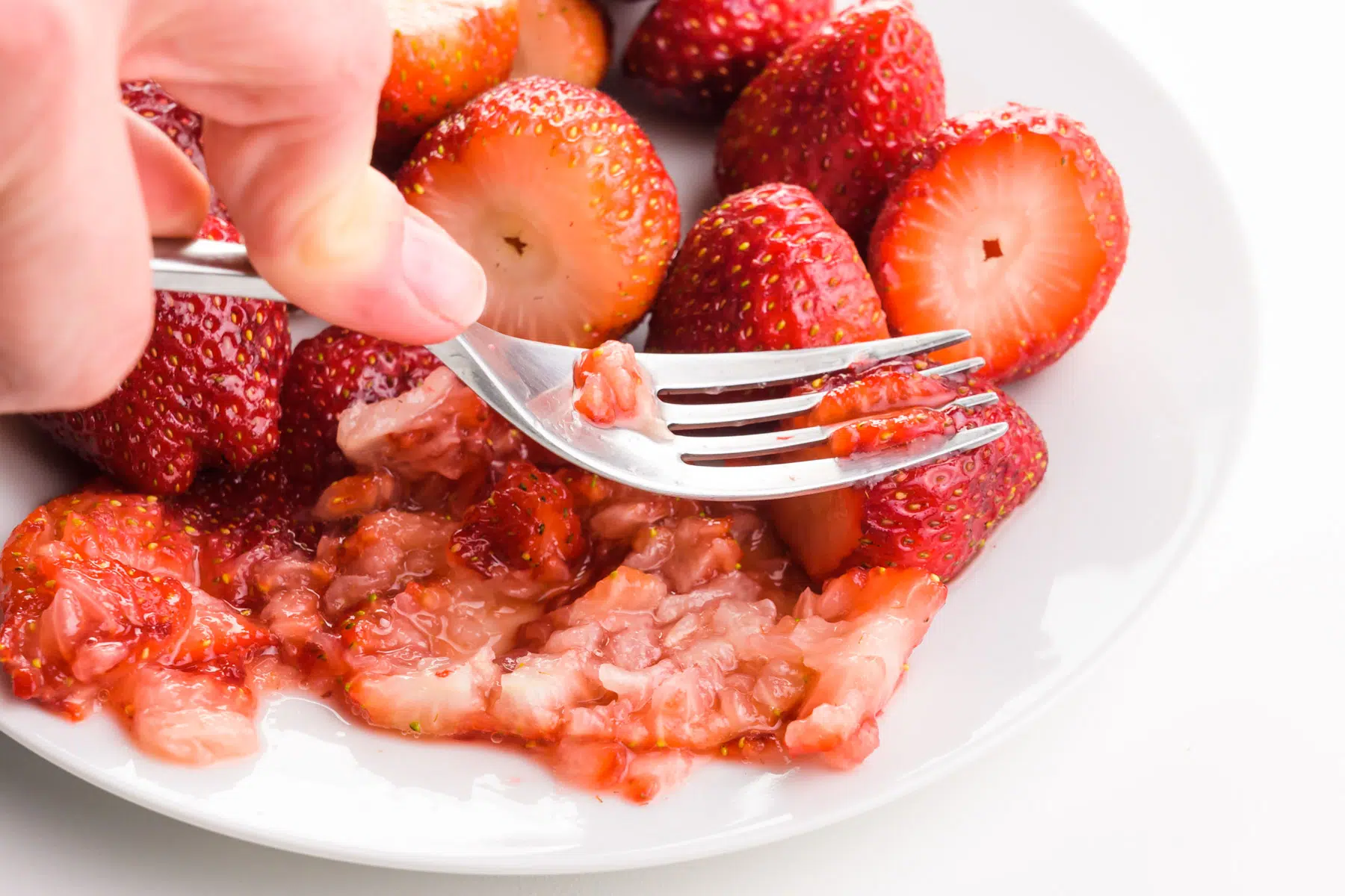 A hand holds a fork using it to mash fresh strawberries.