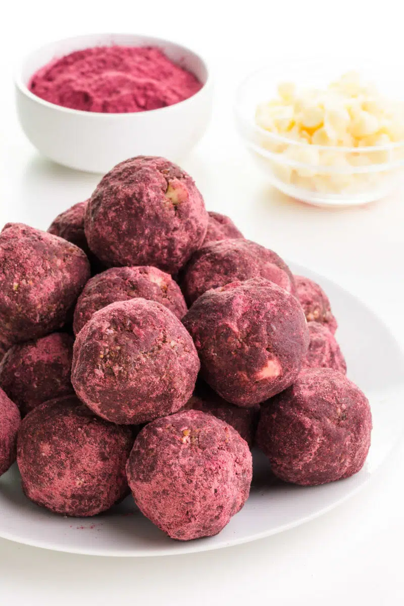 A stack of red velvet protein balls sits in front of a bowl of hibiscus powder and white chocolate chips.