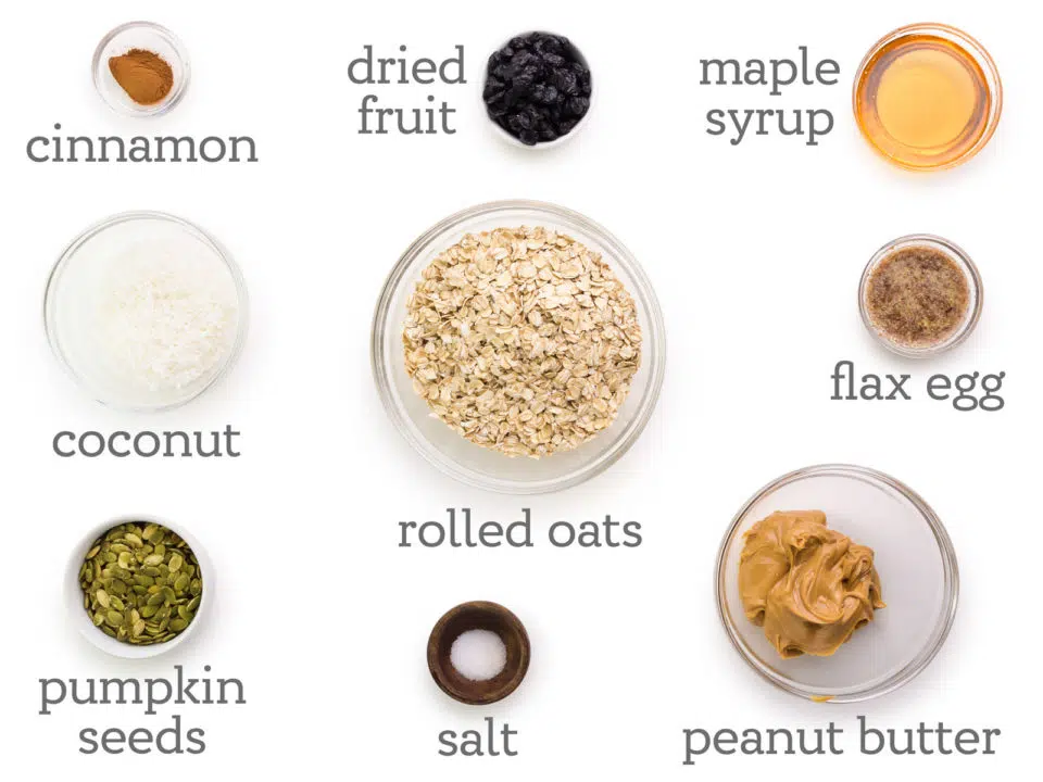 Ingredients are laid out on a white counter. The labels next to them ready, dried fruit, maple syrup, flax egg, peanut butter, salt, rolled oats, pumpkin seeds, coconut, and ground cinnamon.