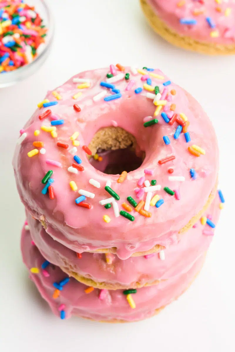 Looking down on a stack of gluten-free donuts with pink icing and sprinkles. There are more donuts in the background.
