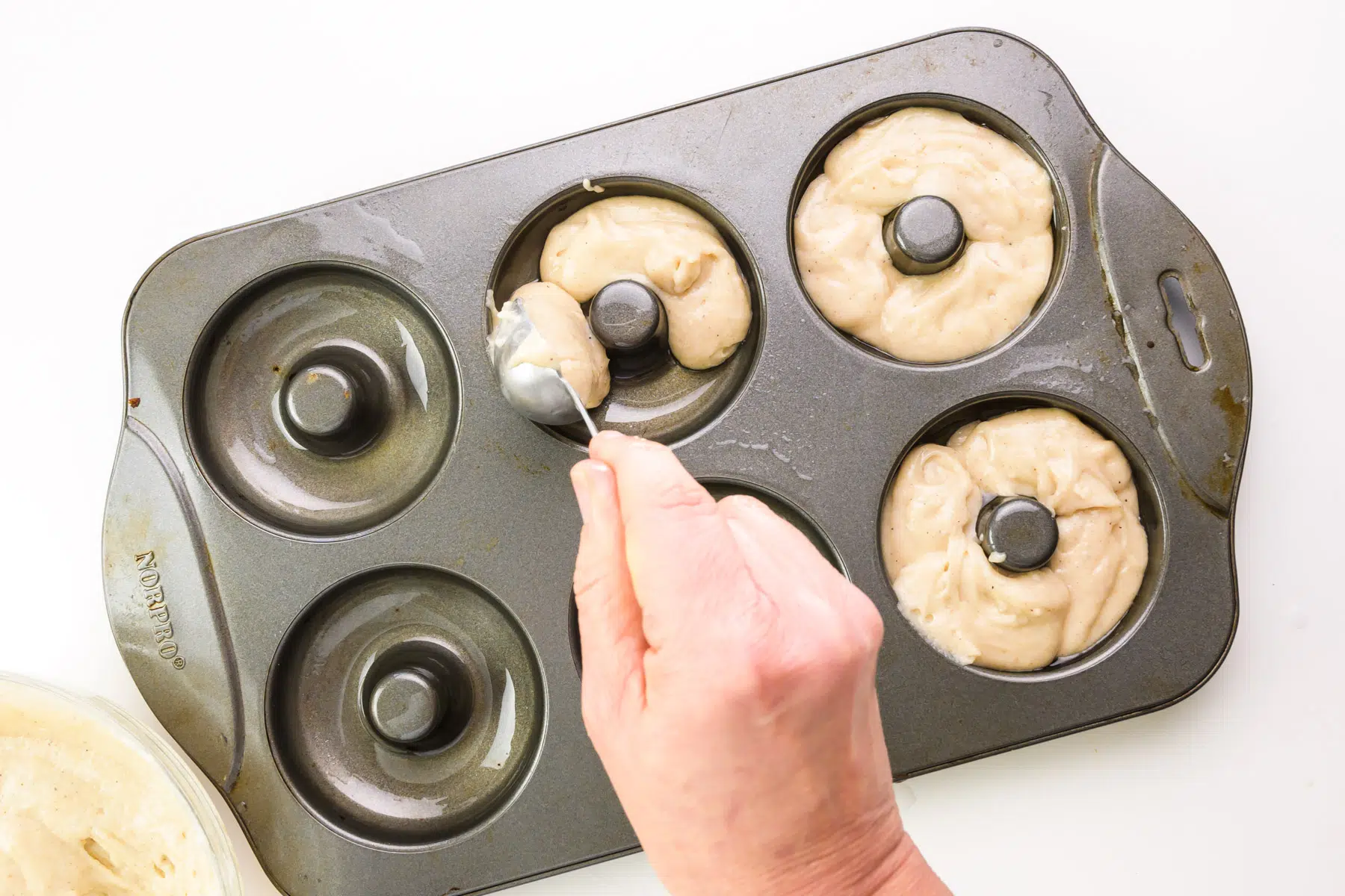 A hand holds a spoon, dispensing batter into greased donut compartments of a baking pan.