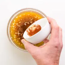A hand holds a bowl of spices adding them to another bowl with a broth mixture.