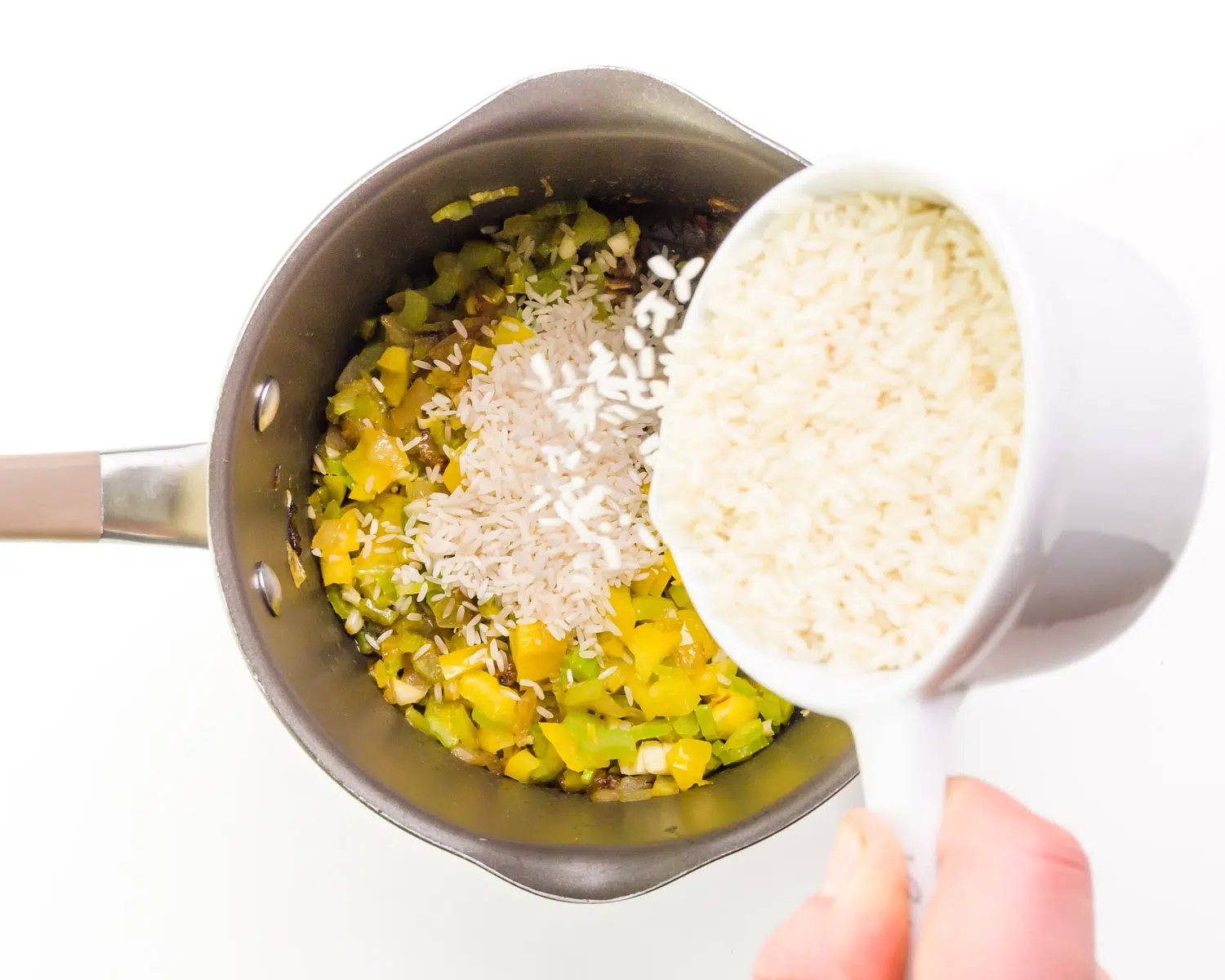 A hand holds a measuring cup of rice pouring it into a saucepan with cooked aromatic vegetables.