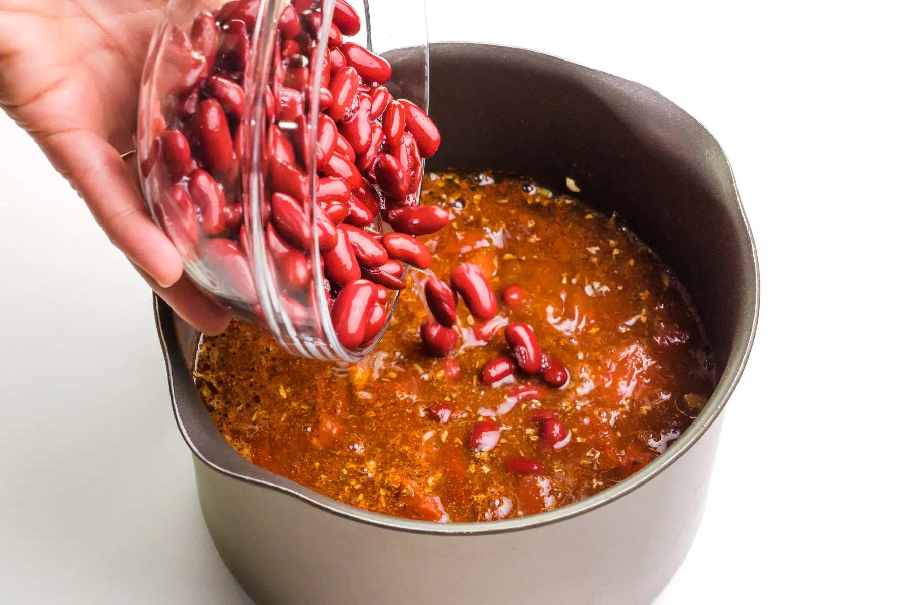 Kidney beans are being poured into a saucepan with tomato-based sauce.