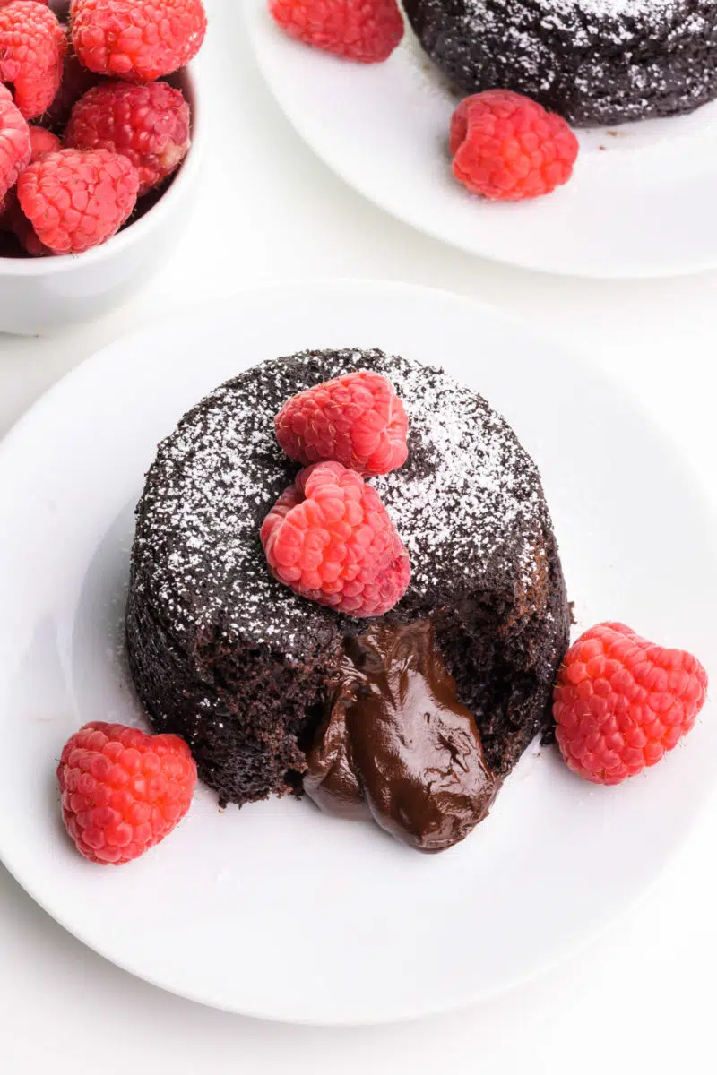 Looking down on a lava cake with melted chocolate pouring out. It has fresh raspberries on top. There's another plate and a bowl of raspberries in the background.