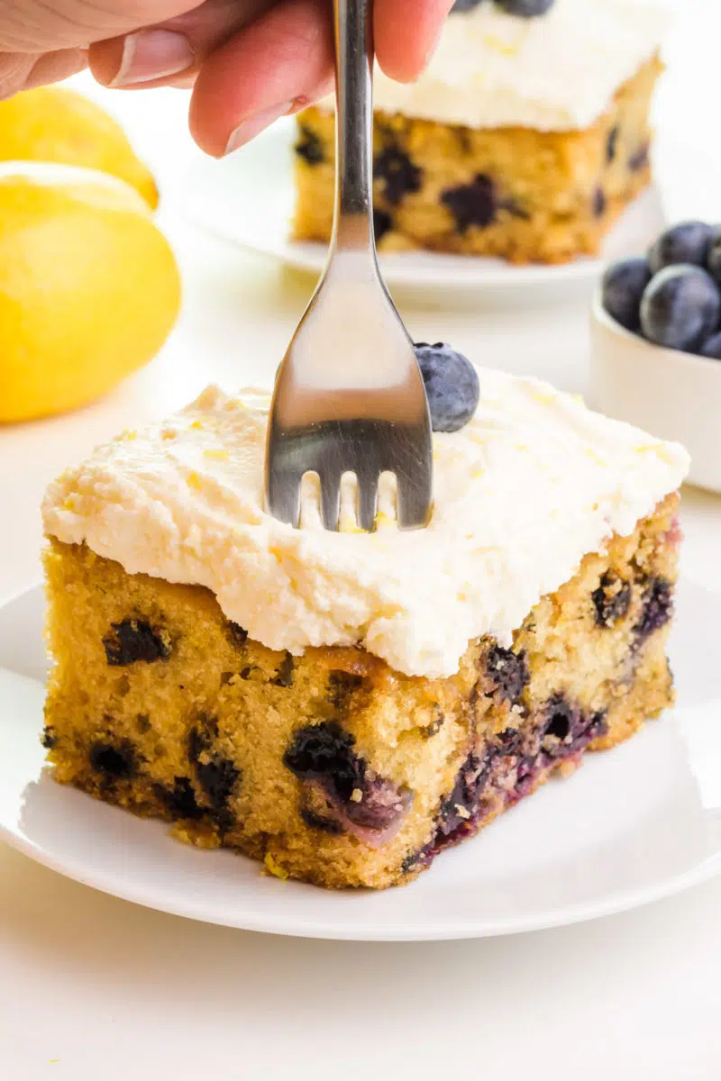 A hand pushes a fork into a slice of lemon blueberry cake. There are fresh lemons, blueberries, and another slice of cake in the background.