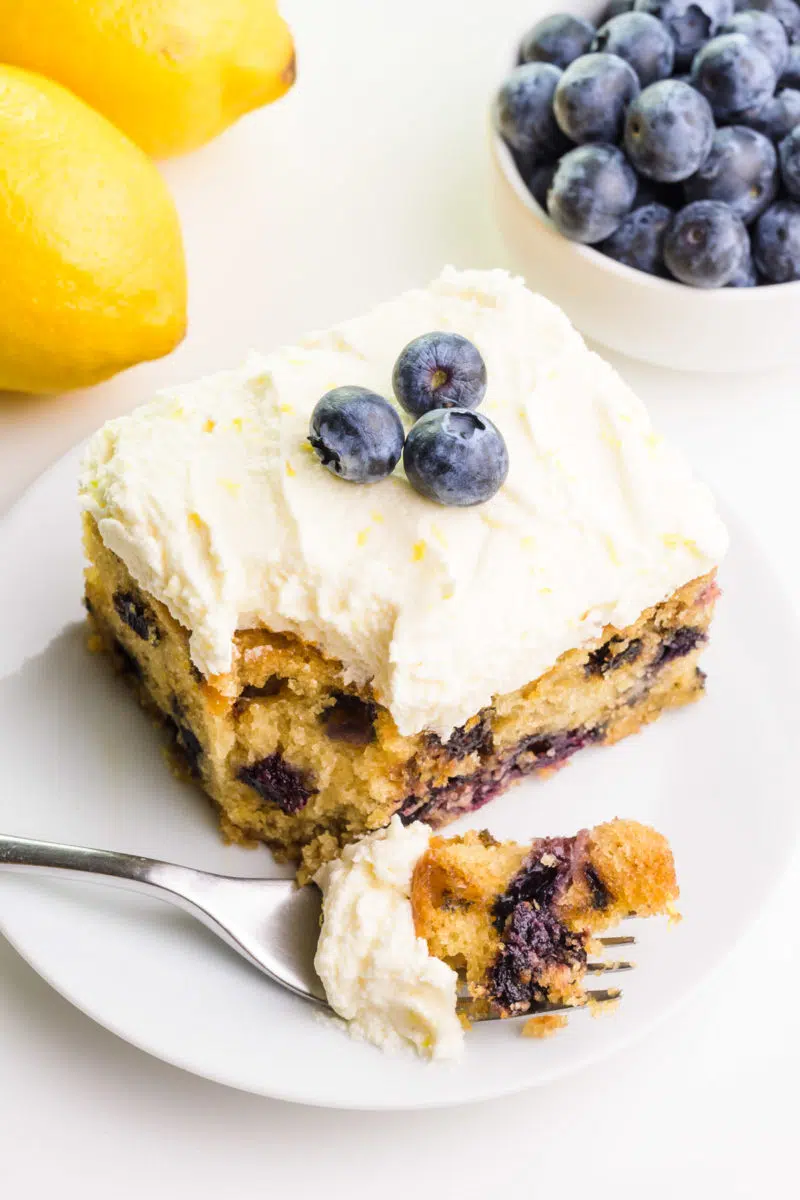 Looking down on a slice of vegan lemon blueberry cake with a bite taken out. There are fresh blueberries and lemons in the background.