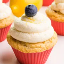 A cupcake has vegan lemon frosting on top with a blueberry and lemon zest. There are more cupcakes and a lemon in the background.