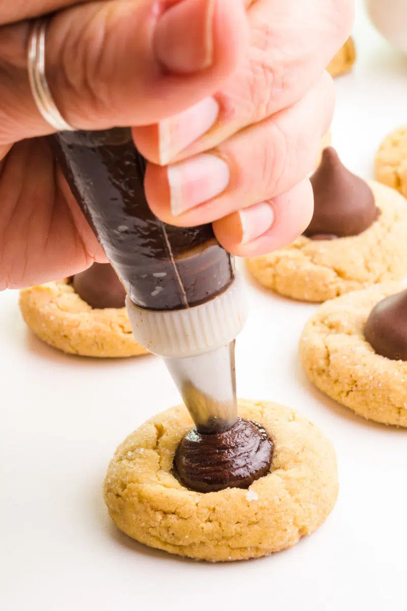 A hand holds a piping bag, pressing chocolate ganache into the center of a cookie.
