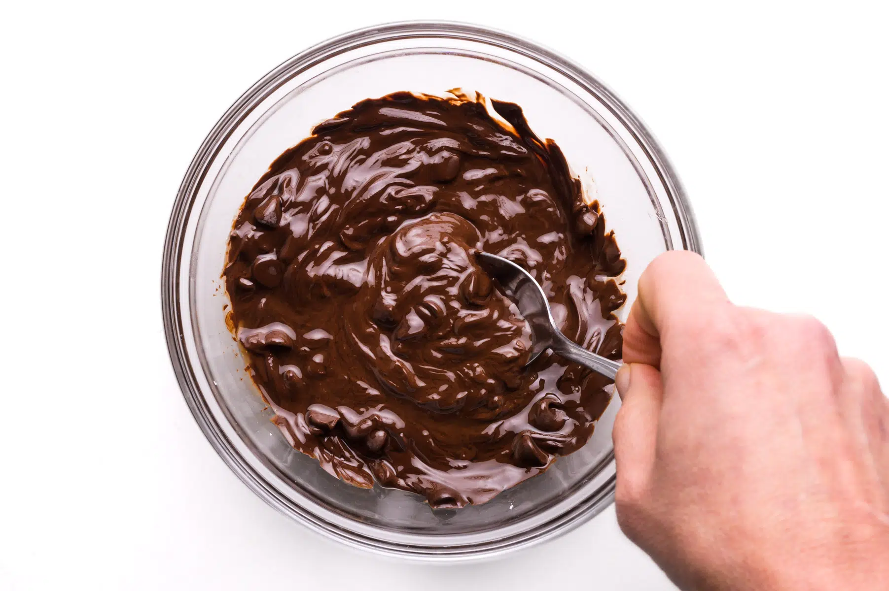 A hand holds a spoon stirring melting chocolate chips in a bowl.