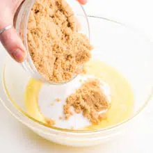 A hand holds a bowl of brown sugar, pouring it into a bowl with melted butter and granulated sugar.