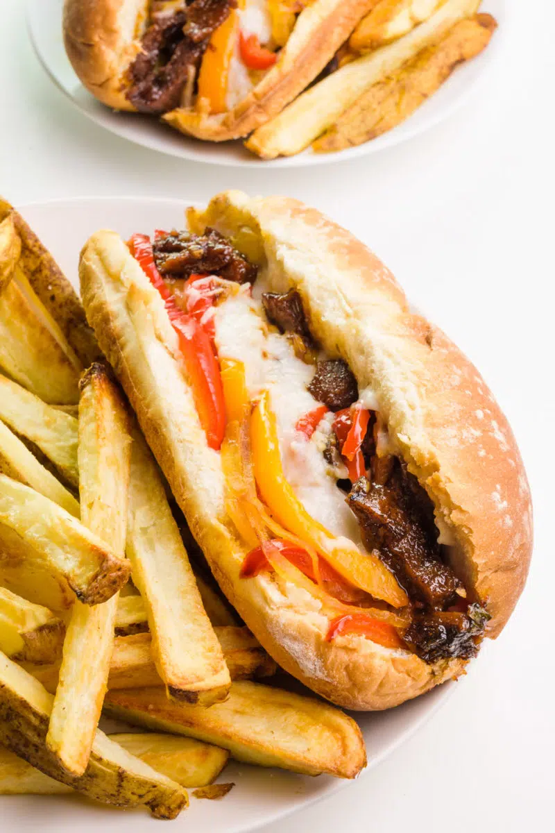 A vegan Philly cheesesteak sandwich sits on a plate next to fries. There's another plate in the background.