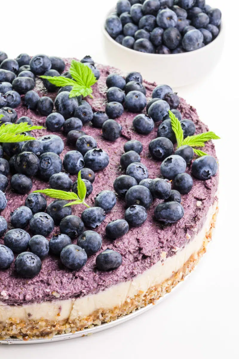 A raw cashew cheesecake has fresh blueberries on to and sits in front of a bowl of fresh blueberries.