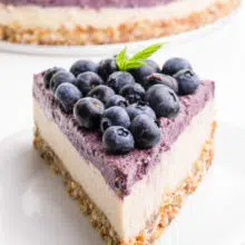A slice of raw cheesecake has blueberries on top. It's sitting on a white plate. The rest of the cheesecake is in the background.
