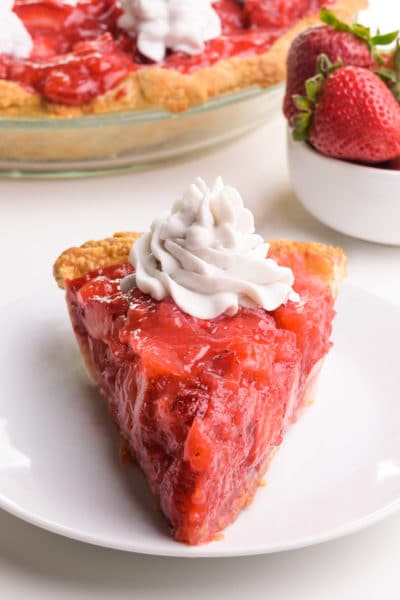 A slice of vegan strawberry pie sits on a plate. It has whipped cream on top. There is more pie and a bowl of strawberries in the background.