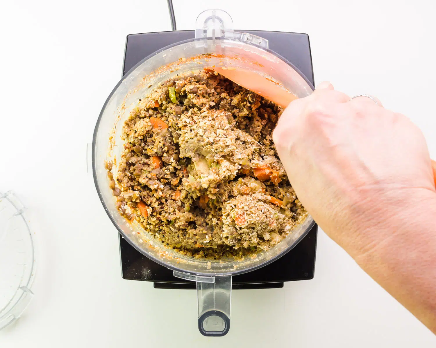 A hand holds a rubber spatula, using it to scrape down the side of a food processor bowl.