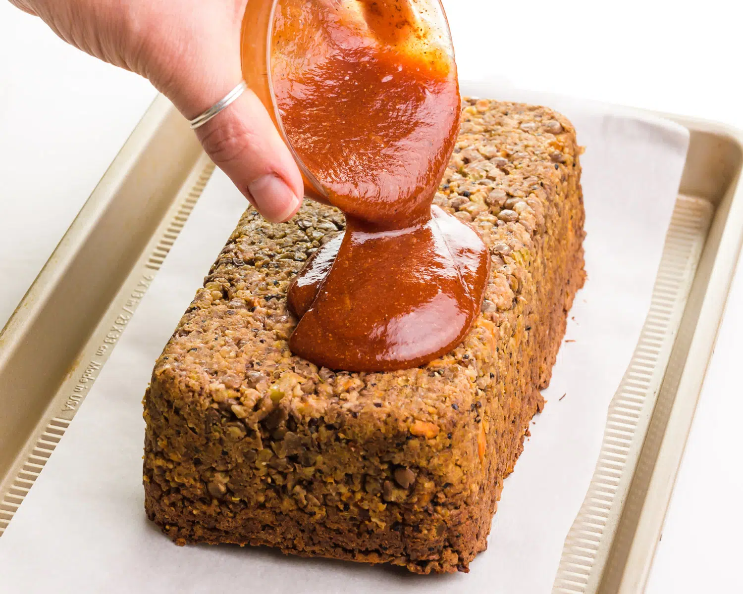 A hand holds a bowl, spreading a ketchup mixture over a bean loaf.