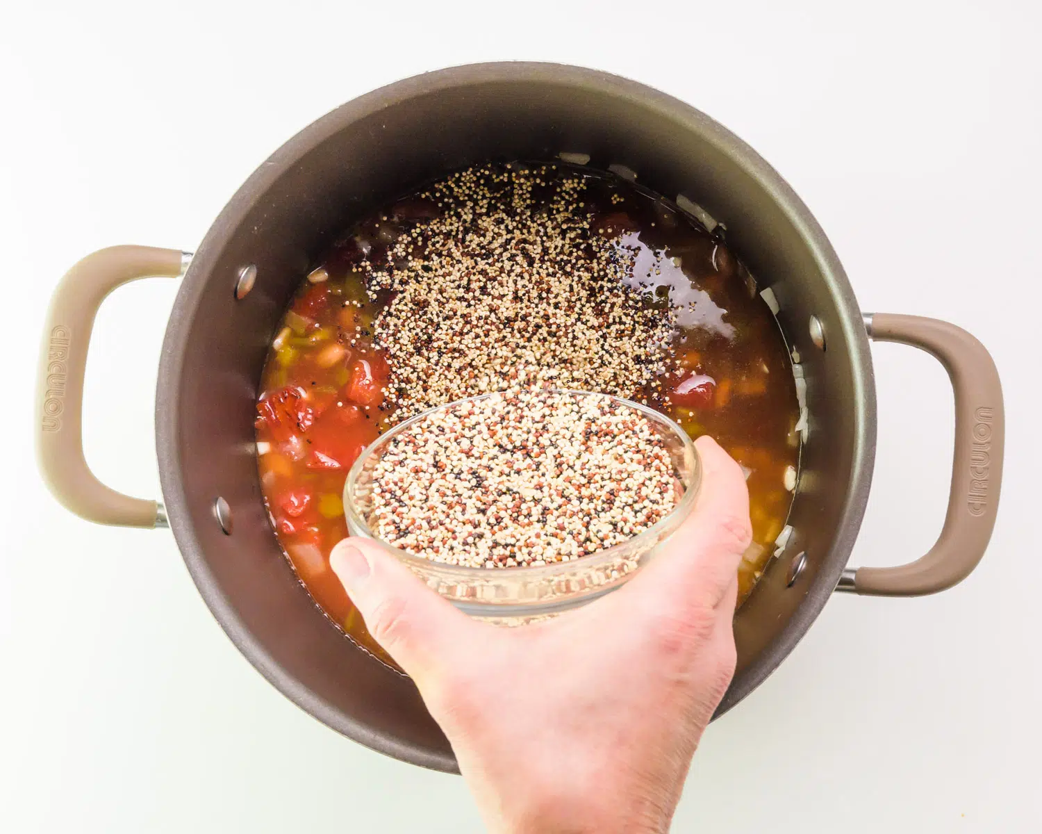 A hand holds a bowl of quinoa, adding it to a large pot with other ingredients.
