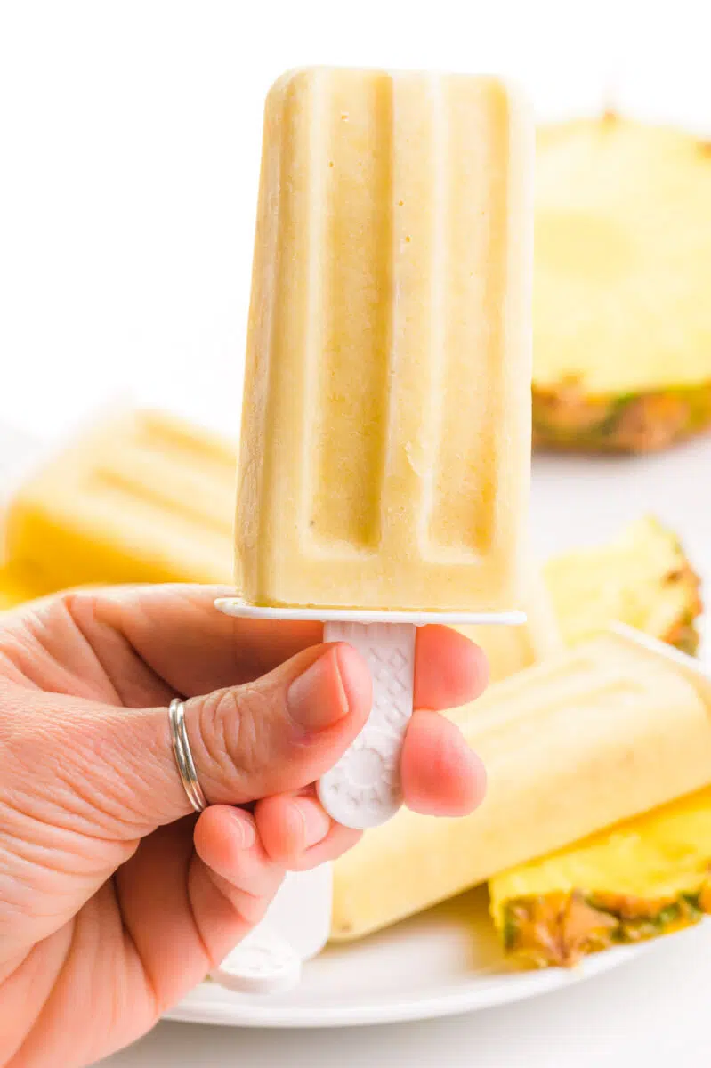 A hand holds a pineapple popsicle in front of a more popsicles and fresh pineapple in the background.