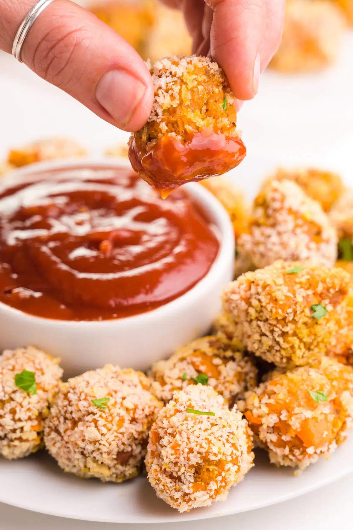 A hand holds a sweet potato  tater tot dipped in ketchup, dangling over a plate with more tater tots and a bowl of ketchup.